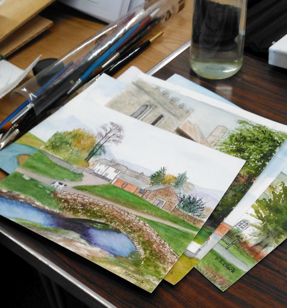A pile of watercolours on a table with brushes and a water pot.