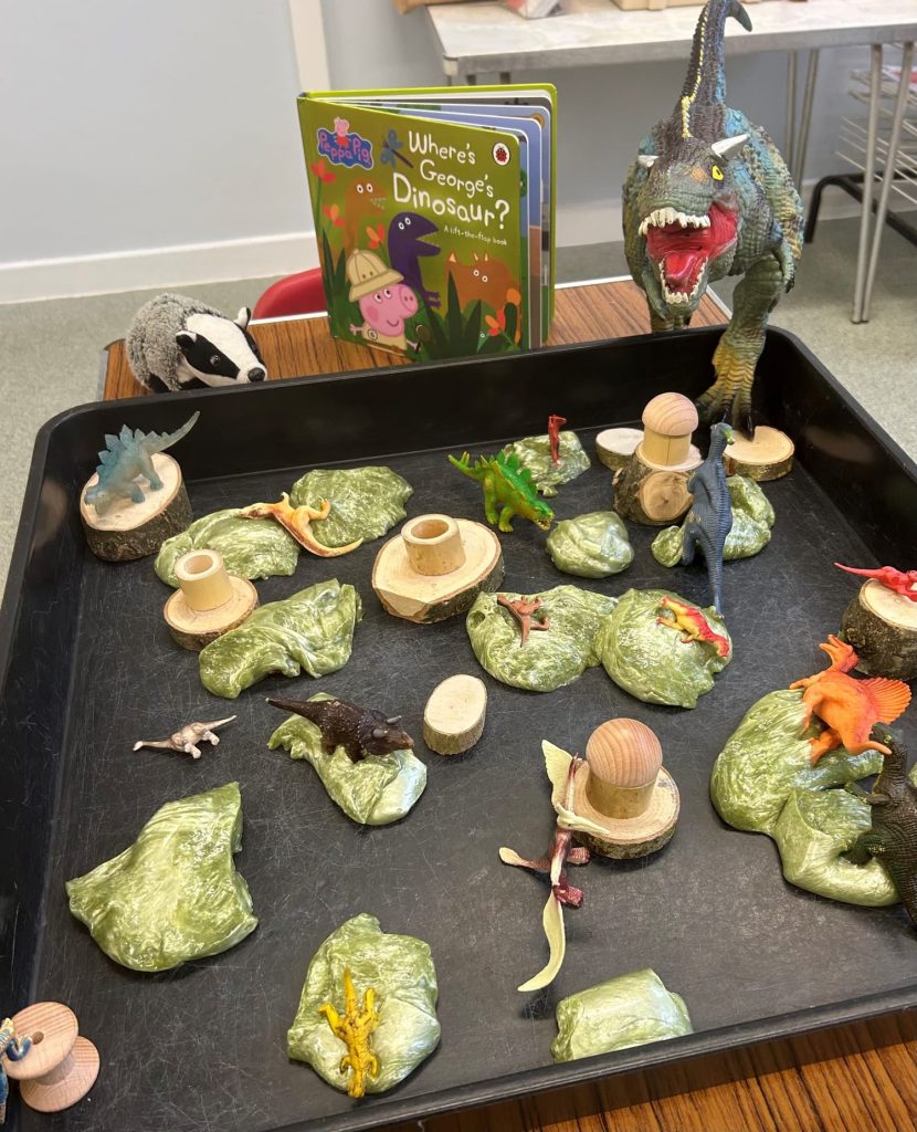 A tray of dinosaur toys next the the book "Where's George's Dinosaur" and a stuffed badger toy called Mr Badger.