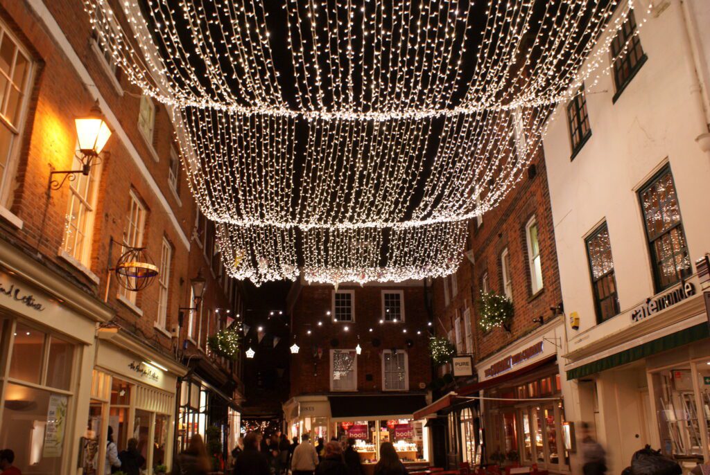 A night-time view of The Square in Winchester, with brick shopfronts and restaurants lit by a canopy of white fairy lights as people walk below.
