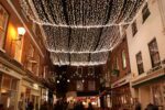 A night-time view of The Square in Winchester, with brick shopfronts and restaurants lit by a canopy of white fairy lights as people walk below.