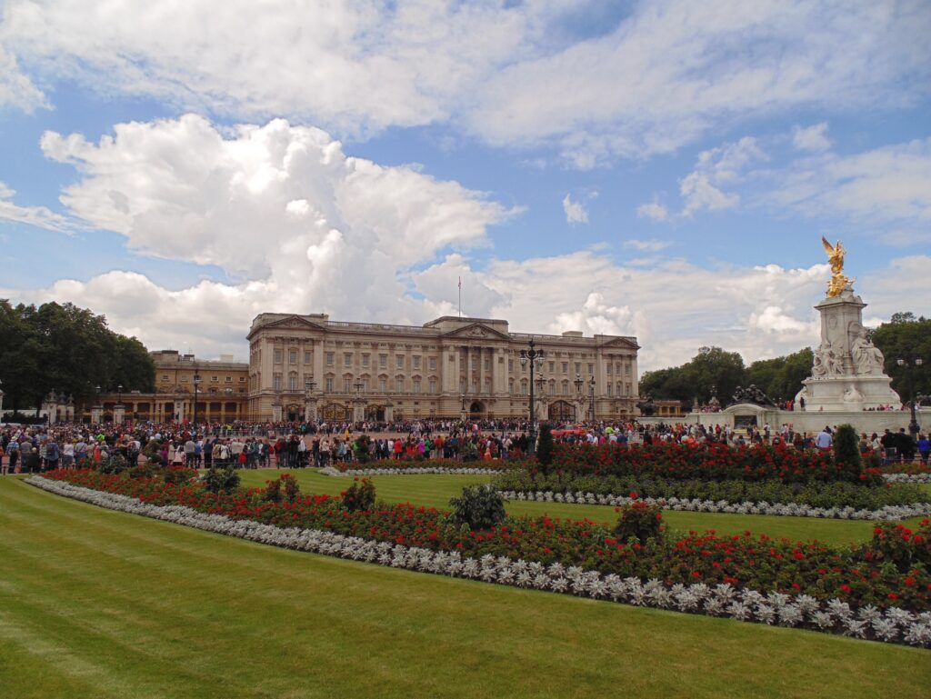 Buckingham Palace with crowds gathered at the gates, formal gardens in the foreground, and the Victoria Memorial to the right under a partly cloudy sky.