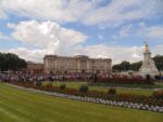 Buckingham Palace with crowds gathered at the gates, formal gardens in the foreground, and the Victoria Memorial to the right under a partly cloudy sky.