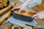 Hands holding a freshly baked loaf in a tin on a striped cloth, surrounded by assorted breads including baguettes, croissants and sliced loaves on a wooden surface.