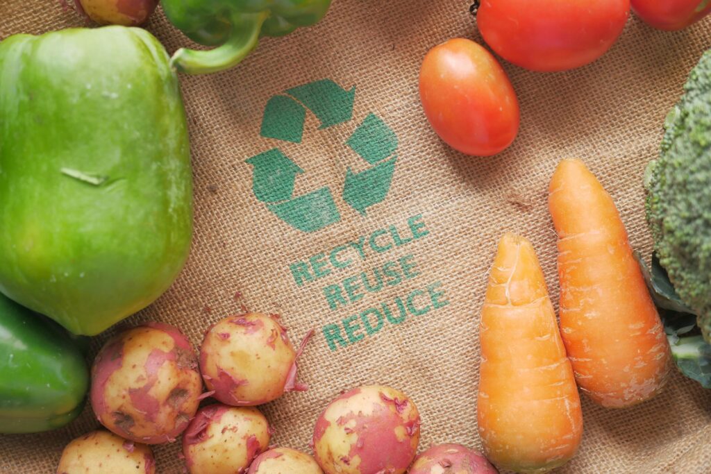 A variety of fresh vegetables — including green peppers, red potatoes, carrots, tomatoes, and broccoli — arranged on burlap fabric with a green recycling symbol and the words “RECYCLE REUSE REDUCE” printed in the centre.