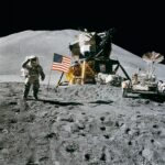 Astronaut on the Moon during Apollo 15, saluting beside the American flag with the lunar module and equipment behind on the grey, dusty surface.